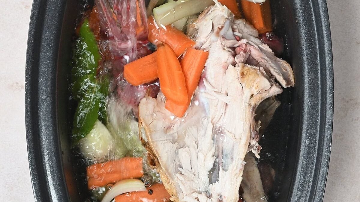 A slow cooker filled with chicken carcass, carrot pieces, onion chunks, green vegetables, and peppercorns as water is poured in—perfect for making chicken stock at home.