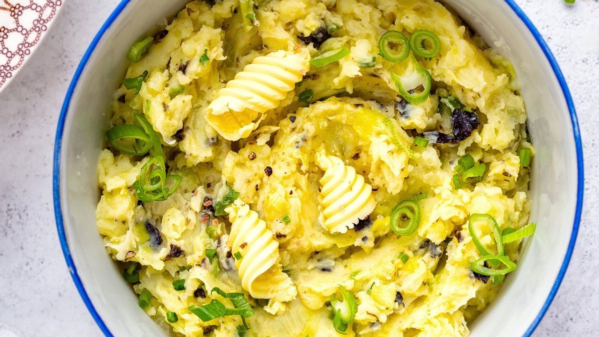 A white pot filled with mashed potatoes, topped with swirls of butter and chopped green onions, on a light surface with a patterned plate and green onions nearby.