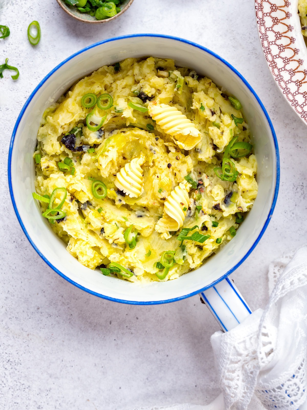 A white pot filled with mashed potatoes, topped with swirls of butter and chopped green onions, on a light surface with a patterned plate and green onions nearby.