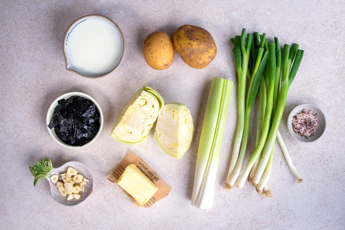 A flat lay of ingredients on a light surface: a pitcher of milk, two potatoes, several spring onions, a bowl of salt, a leek, half a cabbage, a dish of seaweed, sliced garlic, and a pat of butter on wax paper.