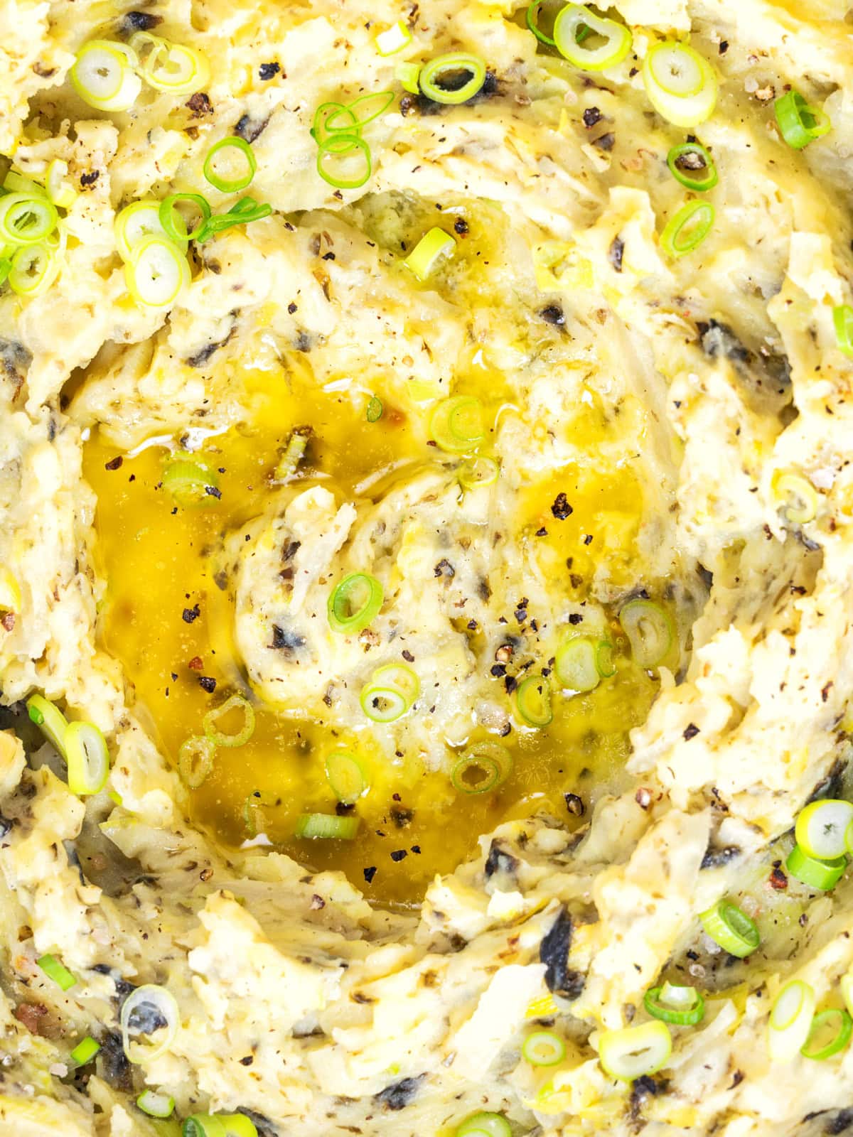 Close-up of creamy colcannon mashed potatoes topped with chopped spring onions, cracked black pepper, and melted butter pooling in the centre. Flecks of seasoning and herbs are visible throughout the dish.