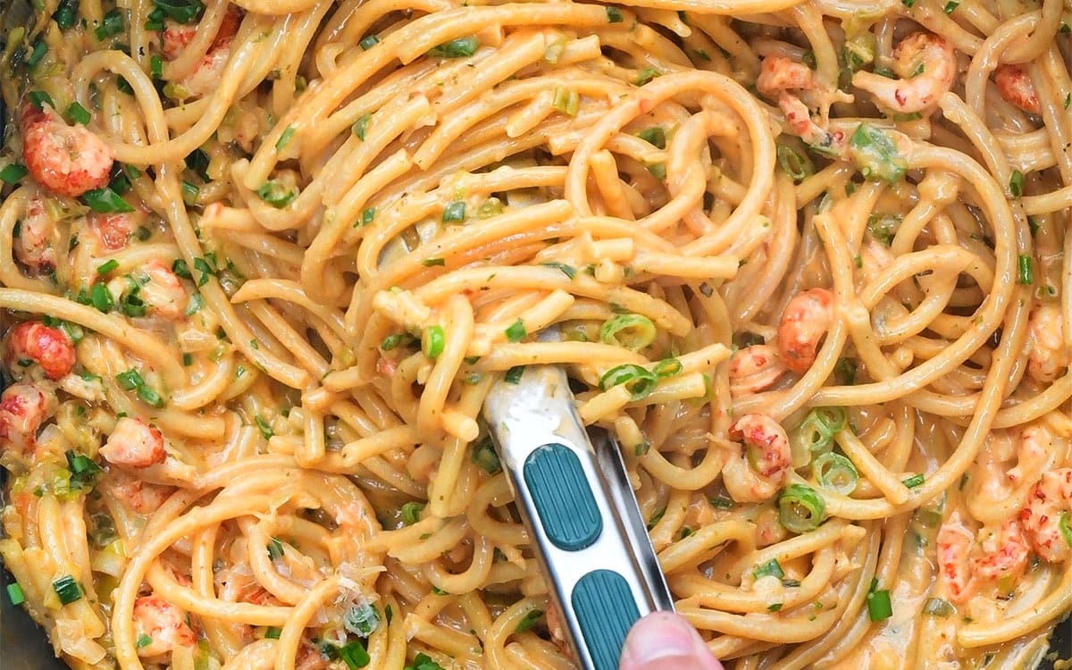 A close-up of creamy crawfish pasta with herbs, chopped spring onions, and pieces of seafood being stirred with tongs.