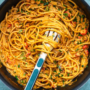 A pan filled with creamy crawfish pasta cooked in one saucepan, mixed with small pieces of seafood and garnished with chopped herbs. A slotted spoon is resting in the pasta, ready to serve.