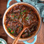 A blue Dutch oven filled with rich, Guinness braised short ribs stew, garnished with fresh herbs, sits on a wooden surface. A wooden spoon rests in the pot, and a light blue cloth is partially visible beside it.