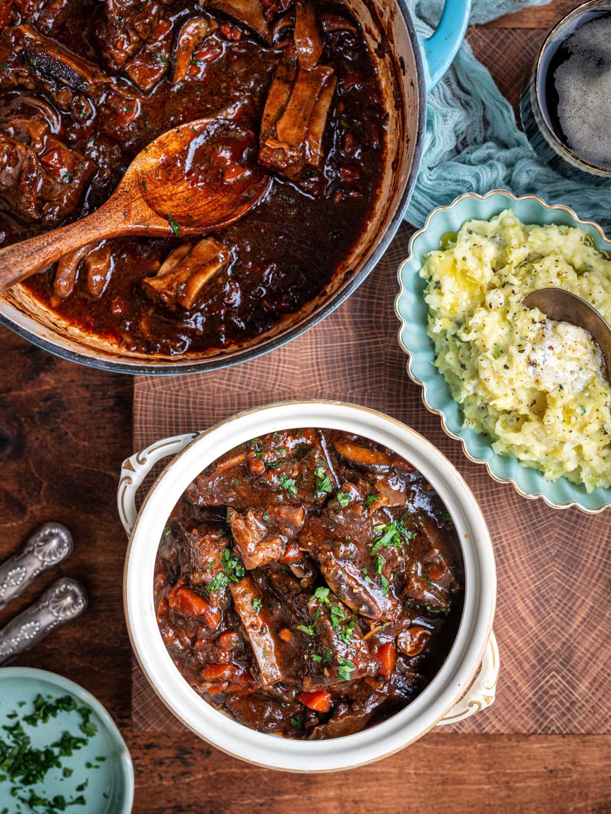 A hearty beef stew with vegetables and tender Guinness short ribs in a white bowl garnished with parsley, beside a pot of stew and creamy mashed potatoes with butter, all set on a wooden table—the ultimate comfort food.
