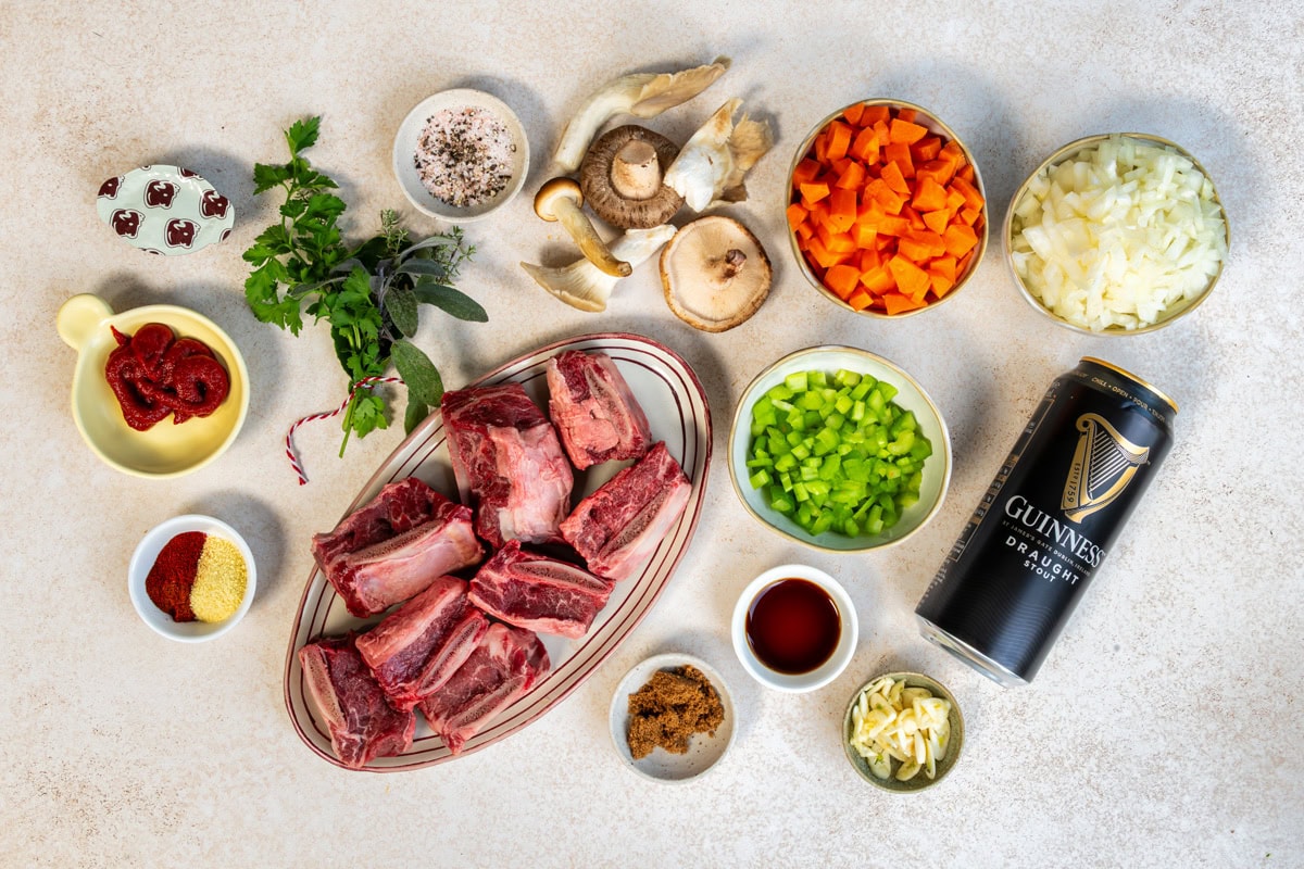 An overhead view of ingredients for a Guinness braised short ribs, including raw beef short ribs, chopped onions, carrots, celery, garlic, mushrooms, tomato paste, herbs, spices, brown sugar, Worcestershire sauce, and a can of Guinness beer.