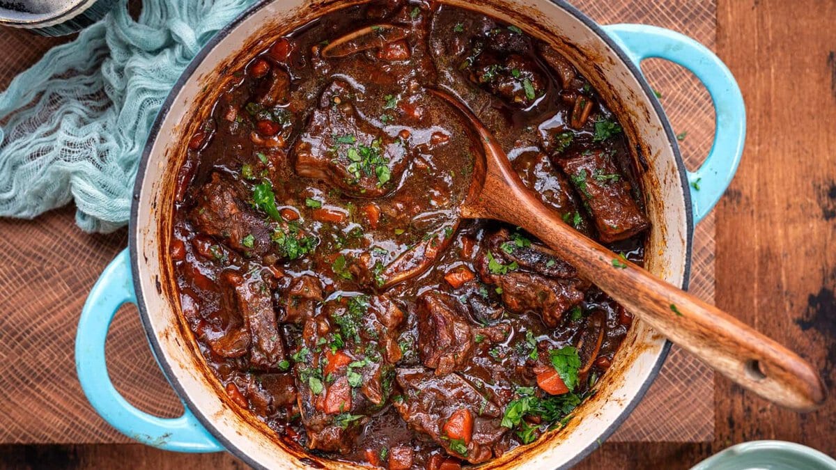 A blue Dutch oven filled with rich, Guinness braised short ribs stew, garnished with fresh herbs, sits on a wooden surface. A wooden spoon rests in the pot, and a light blue cloth is partially visible beside it.
