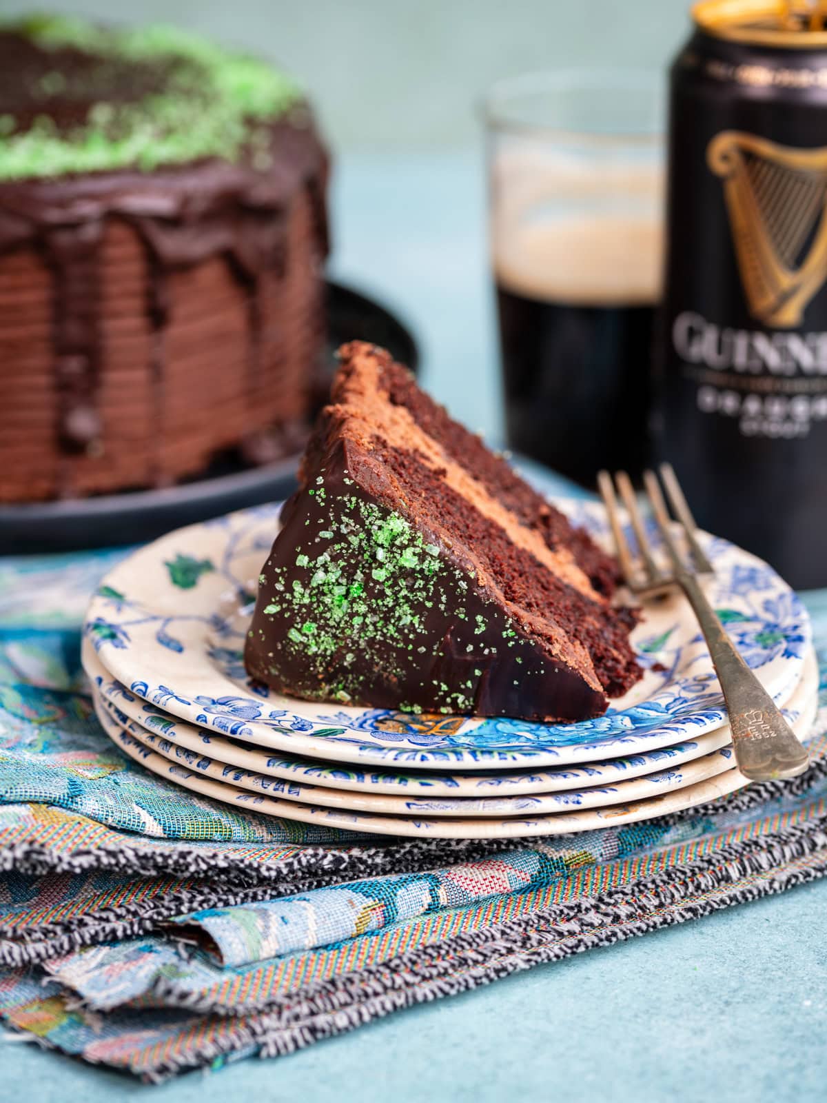 A slice of chocolate stout cake with green sprinkles sits on a stack of floral plates next to a fork, with a can and glass of Guinness and the rest of the cake in the background.