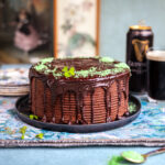 A rich Guinness chocolate cake with dripping chocolate glaze, decorated with green sprinkles and clover leaves, sits on a black plate. A can and glass of Guinness are in the background, along with framed art and stacked plates.