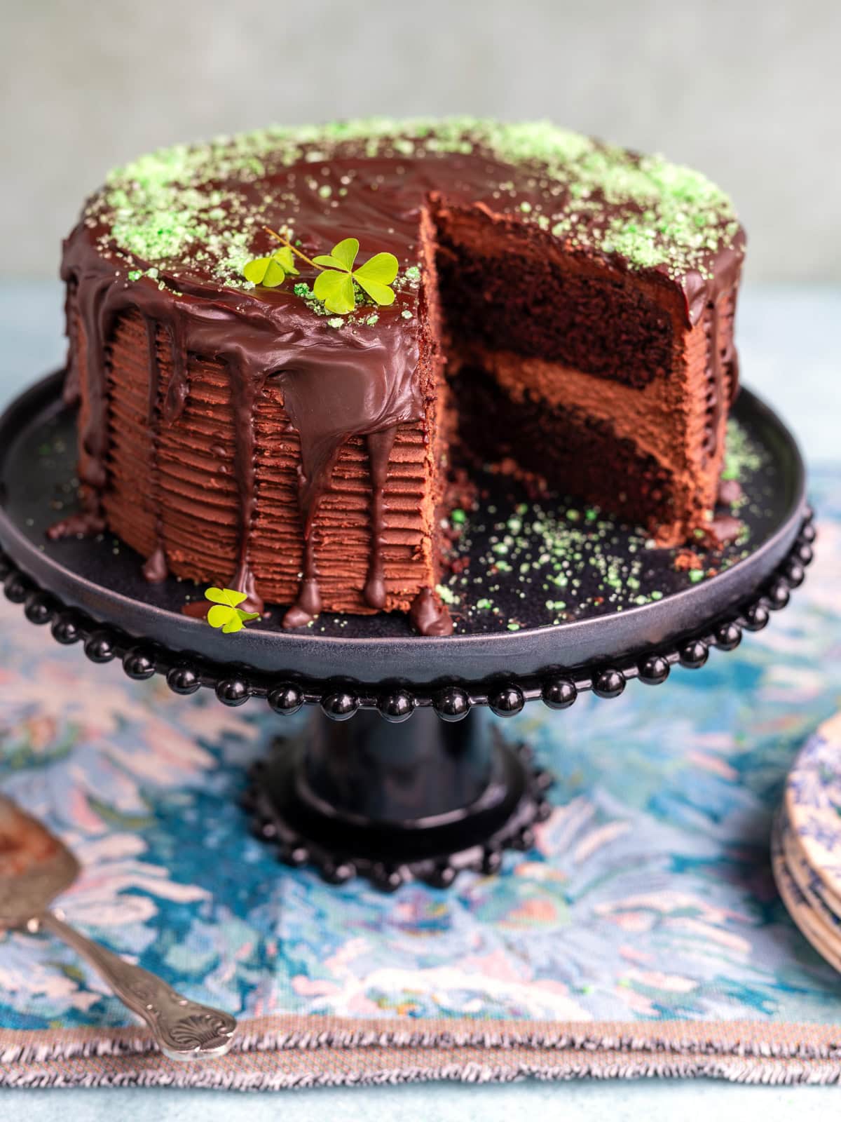 A Guinness chocolate layer cake with chocolate frosting and ganache sits on a black cake stand. The cake is decorated with green sprinkles and clover leaves, and a slice has been cut out to display the layers.