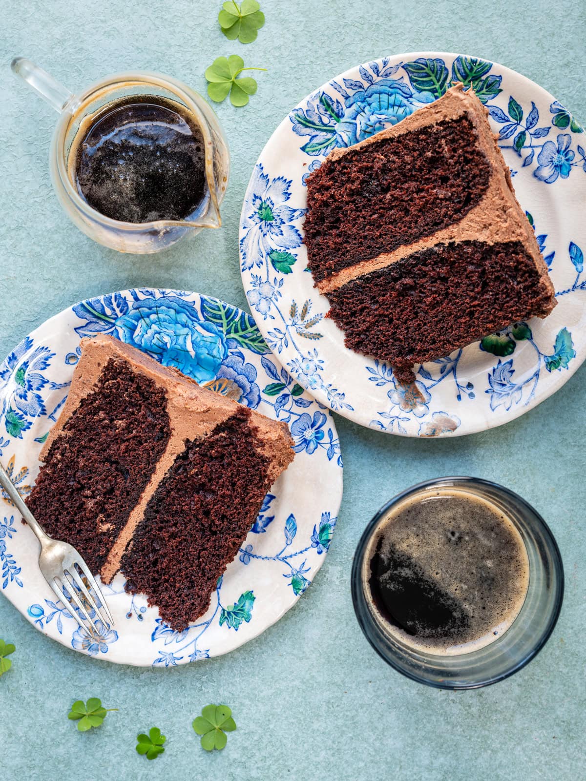 Two slices of chocolate cake with chocolate frosting on floral plates, served with glasses of dark coffee or stout, on a light blue textured surface with a few small clover leaves scattered around.