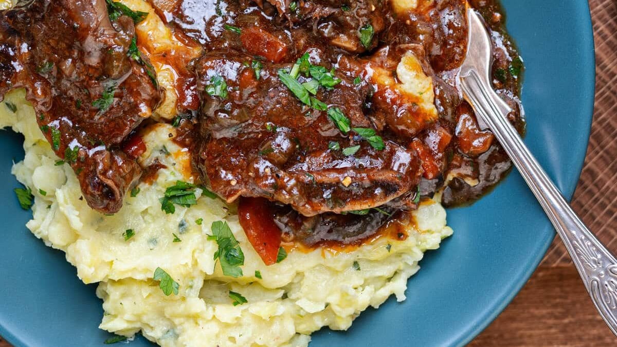 A blue plate with creamy mashed potatoes topped with slow-cooked beef short ribs in a rich, dark Guinness gravy with carrots and herbs, garnished with chopped parsley. A fork rests on the side of the plate.