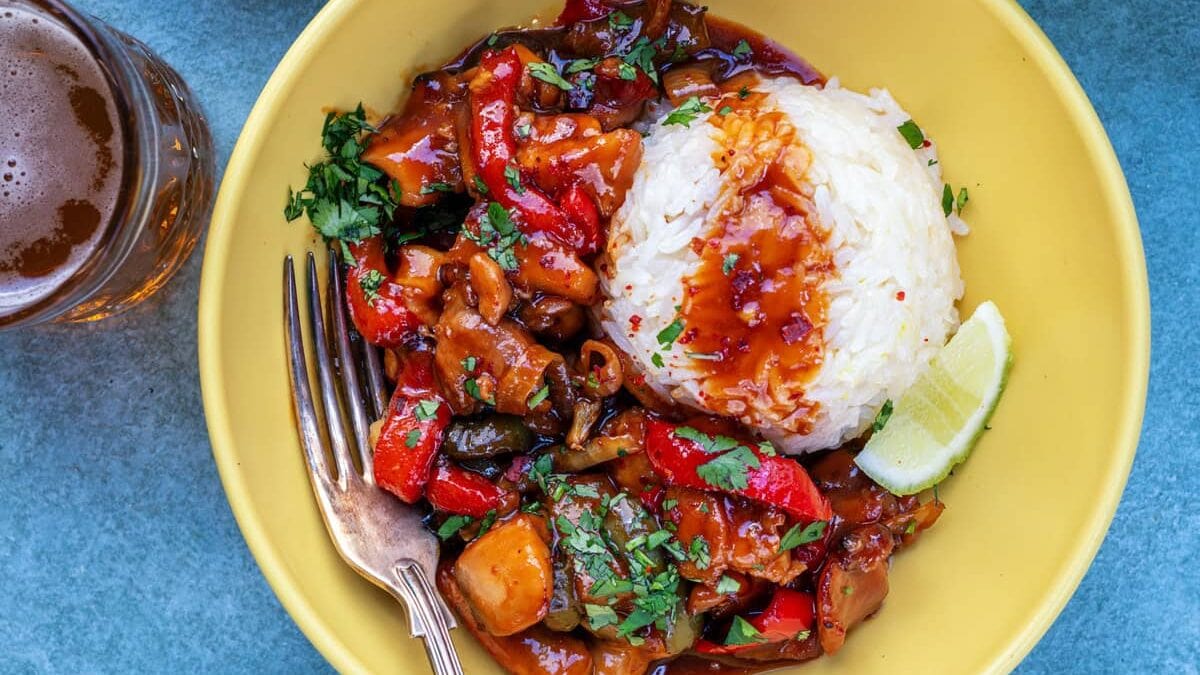 A yellow bowl filled with white rice and a colorful stir-fry of chicken, red peppers, and herbs, garnished with a lime wedge. Nearby are a bowl of rice, a bowl of stir-fry, and a glass of bubbly drink on a blue surface.