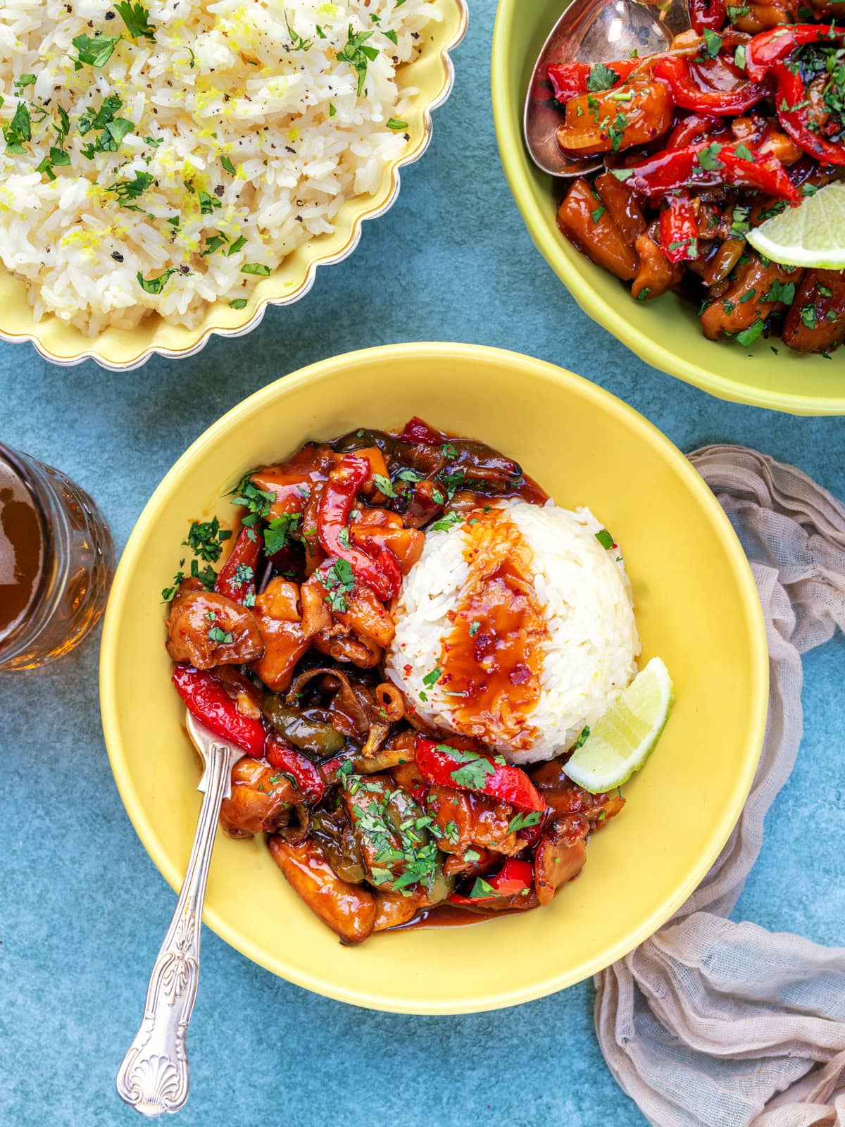 A yellow bowl with pineapple chicken, red bell peppers, and vegetables in sauce, served with a mound of white rice and a lime wedge. Nearby are a bowl of rice, another dish of stir-fry, and a drink on a blue surface.