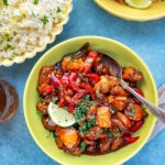 A bowl of Hawaiian pineapple chicken with colorful bell peppers and sauce, garnished with chopped herbs and a lime slice, sits next to a bowl of rice and a small dish of sauce on a blue surface.