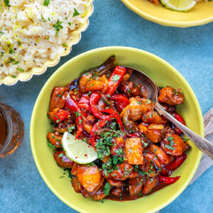 A bowl of Hawaiian pineapple chicken with colorful bell peppers and sauce, garnished with chopped herbs and a lime slice, sits next to a bowl of rice and a small dish of sauce on a blue surface.