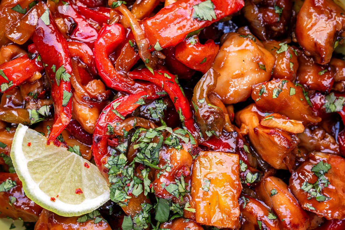 Close-up of a colorful stir-fry dish with glazed chicken, red bell peppers, chopped cilantro, and a slice of lime, all coated in a glossy sauce.