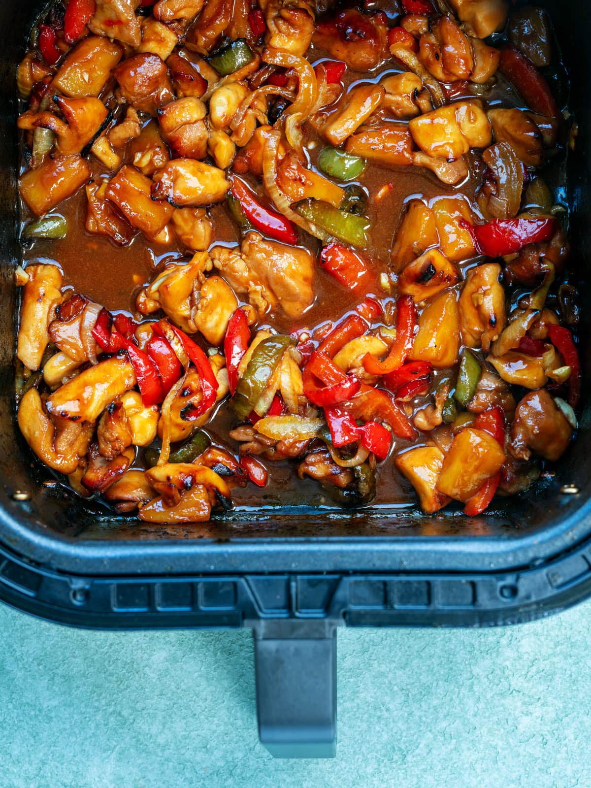 A close-up of cooked sweet and sour chicken with bell peppers and pineapple chunks in a rectangular black air fryer basket on a light surface.
