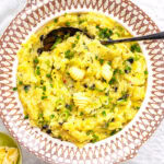 A decorative bowl filled with Irish colcannon—creamy mashed potatoes blended with greens, garnished with chopped chives, black pepper, and butter curls. A serving spoon rests inside, with a small dish of extra butter beside the bowl.