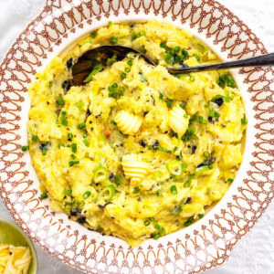 A decorative bowl filled with Irish colcannon—creamy mashed potatoes blended with greens, garnished with chopped chives, black pepper, and butter curls. A serving spoon rests inside, with a small dish of extra butter beside the bowl.