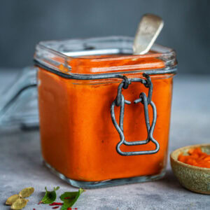 A square glass jar filled with bright orange Onion Tomato Masala sauce, sealed with a metal clasp, sits on a grey surface. A spoon rests inside the jar. Some leaves, spices, and a small bowl are nearby.