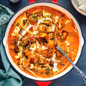 A red-handled frying pan filled with creamy paneer butter masala curry garnished with cream, herbs, and cashew nuts. A serving spoon rests inside. Nearby are a green cloth, a small jug of cream, and a bowl of rice.