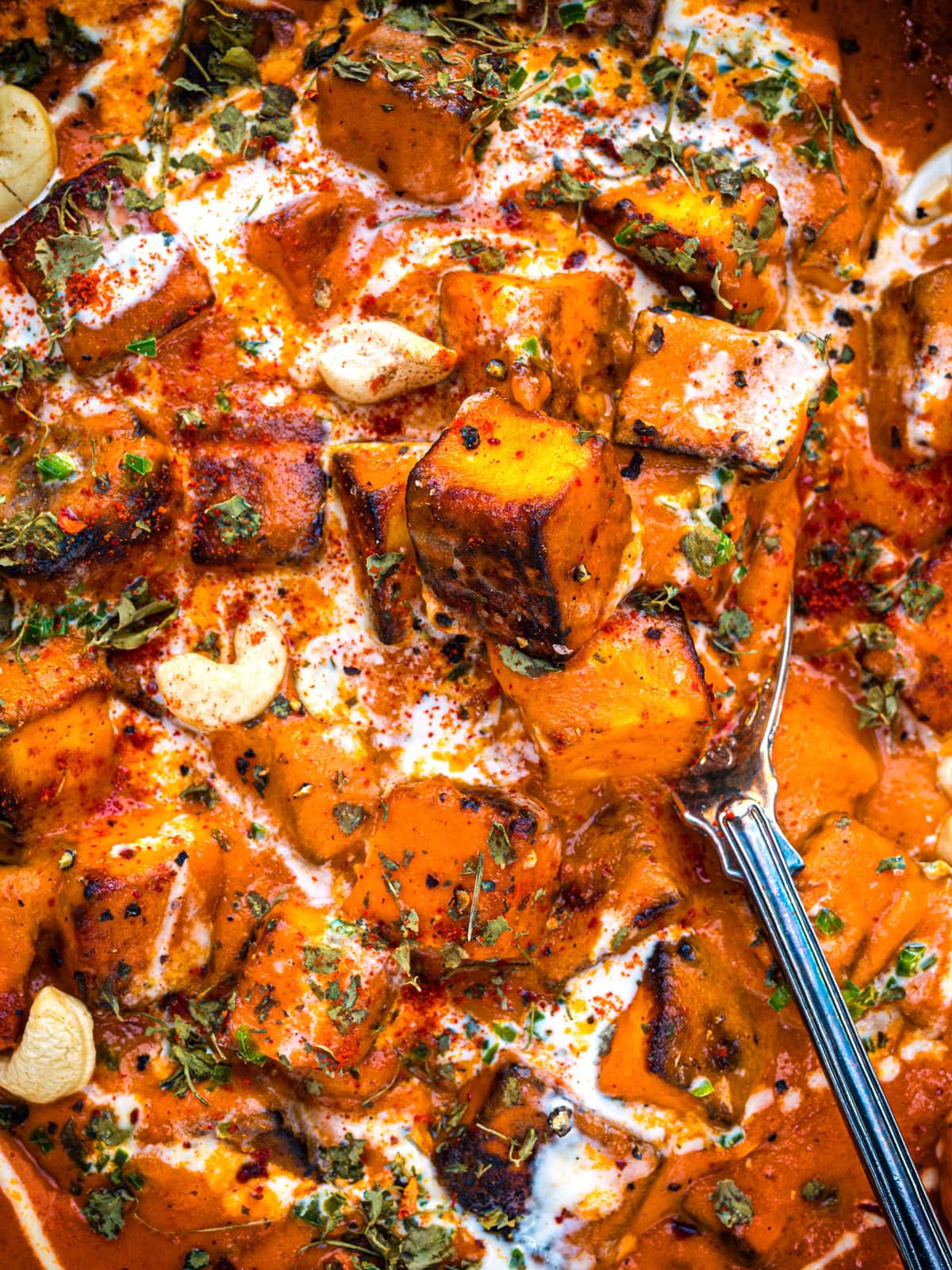 Close-up of creamy, orange-hued roasted paneer and sauce, garnished with herbs, cashew nuts, and a swirl of cream. A metal spoon is visible among the chunky pieces in the rich, spiced dish.