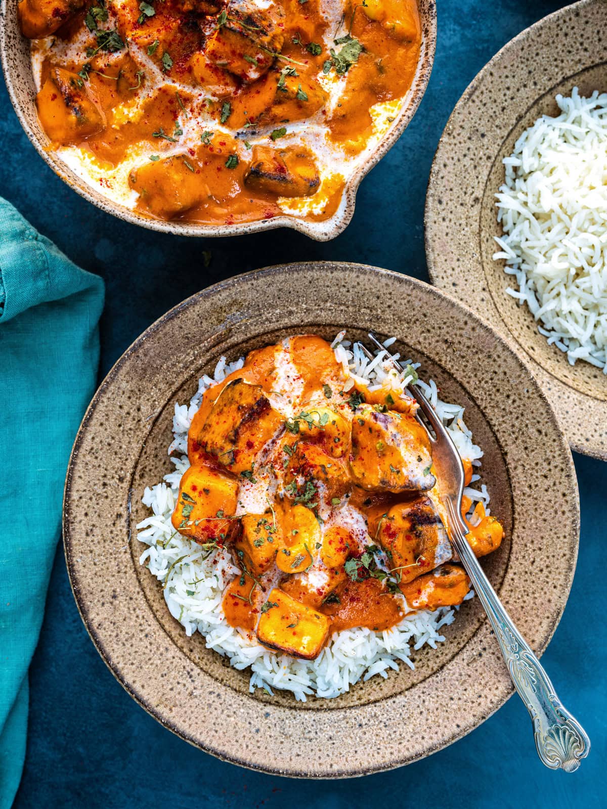 A bowl of white rice topped with paneer makhani curry garnished with cream and herbs, sits next to another bowl of rice and a serving dish of curry on a dark blue surface. A fork rests in the bowl.