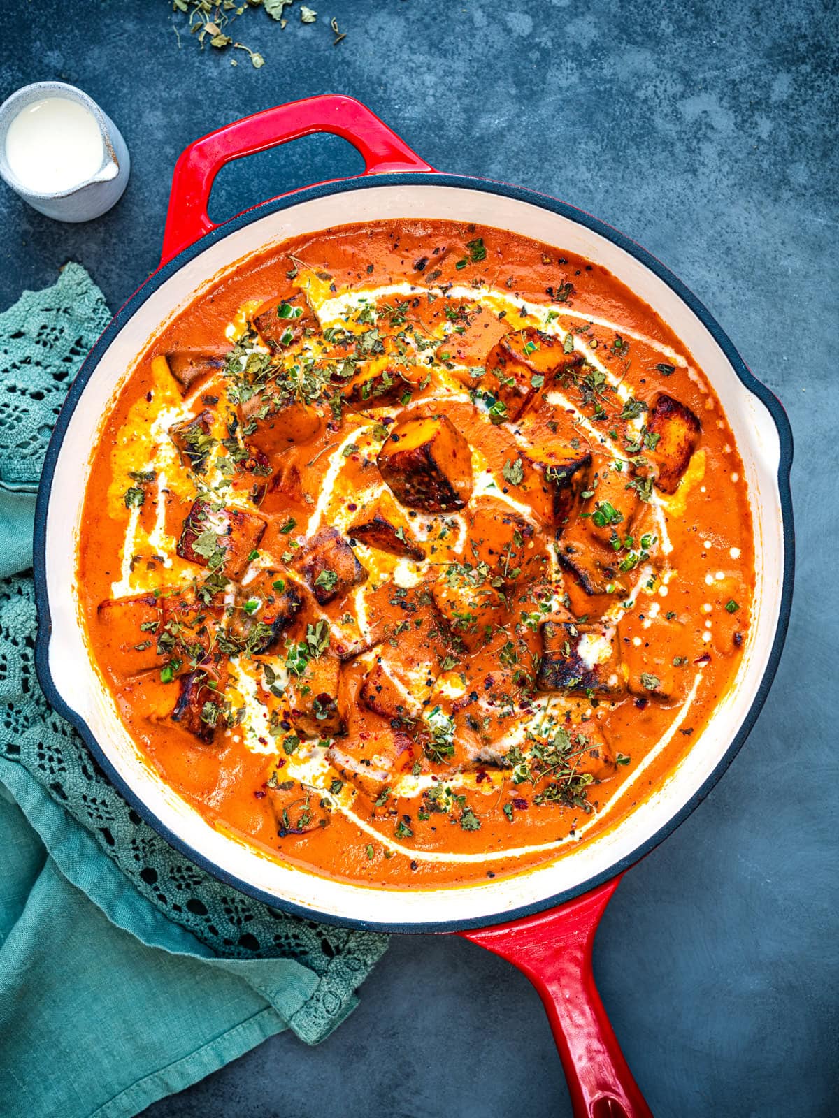 A red pan filled with creamy orange Paneer Butter Masala curry, topped with grilled paneer cubes, drizzled with cream, and garnished with herbs. A green cloth serviette and a small pot of cream sit nearby on a dark surface.