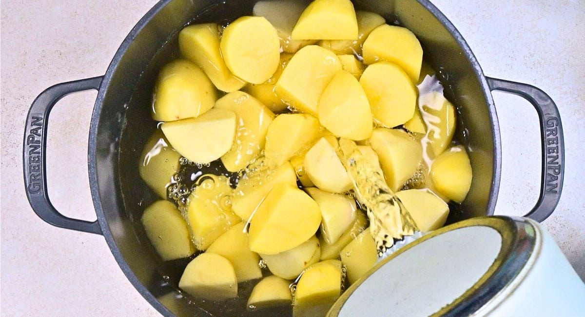 A pot filled with peeled and chopped potatoes for a classic Colcannon recipe is being covered with water poured from a kettle. The pot rests on a light-colored countertop, ready for the next step.