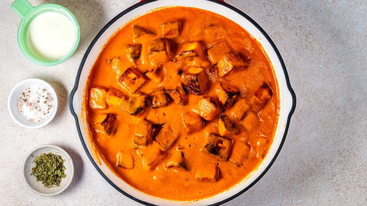 A pan filled with creamy butter paneer curry containing chunks of paneer. Next to the pan are small bowls with milk, dried herbs, and ground spices, all arranged on a light worktop.