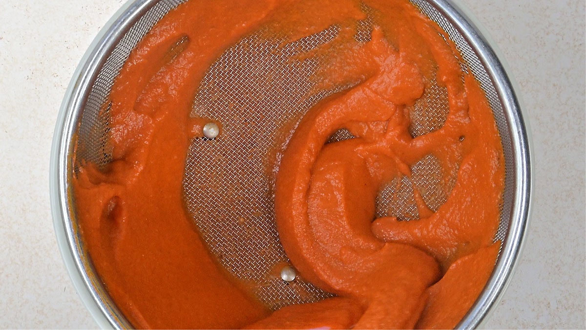 Masala sauce being strained through a metal mesh sieve, with smooth red sauce spread across the sieve’s surface.