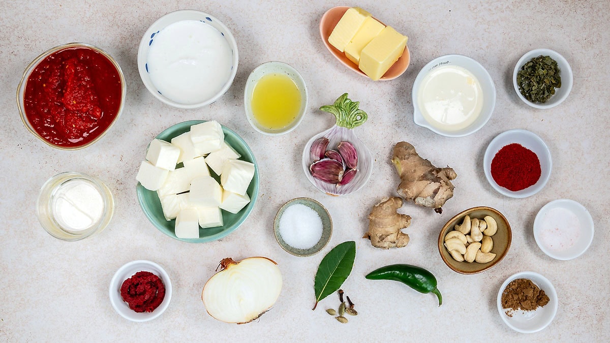 A top-down view of various cooking ingredients arranged on a light surface, including butter, yoghurt, tomato sauce, cream, cubed paneer, spices, cashew nuts, ginger, onion, garlic, green chilli, and small bowls with powders and paste.