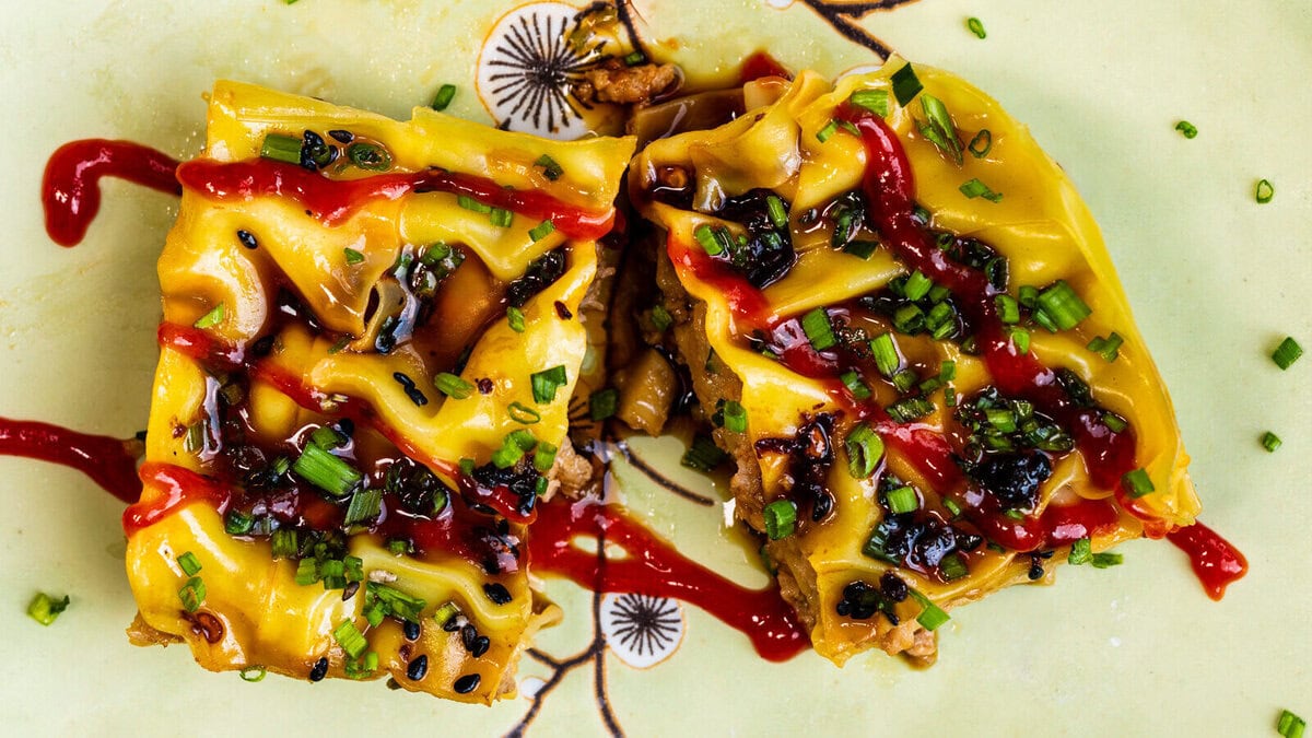 Two square pieces of a savoury dish topped with melted cheese, chopped chives, dark sauce, and zigzag drizzles of red sauce, served on a light yellow plate with a floral design.