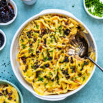 A round baking dish filled with dumpling lasagna, topped with chopped herbs and dark seasoning, sits with a serving spoon and a portion removed. Small bowls of toppings and sauce surround this unique pork dumpling lasagna recipe.