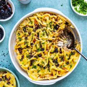 A round baking dish filled with dumpling lasagna, topped with chopped herbs and dark seasoning, sits with a serving spoon and a portion removed. Small bowls of toppings and sauce surround this unique pork dumpling lasagna recipe.