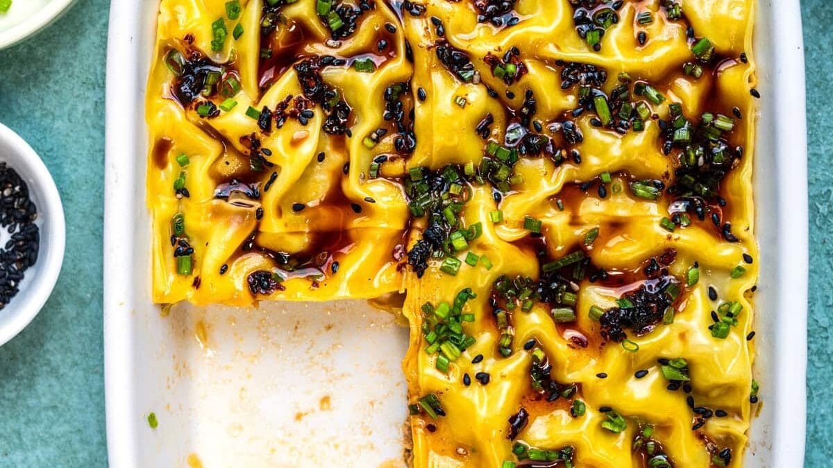 A square white dish holds wavy sheets of wonton wrappers layered with pork filling and topped with chilli oil, soy sauce, and chopped chives. A square portion is missing. Small bowls with chilli oil, chives, and black sesame seeds surround the dish.