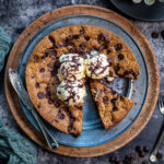 A giant chocolate chip biscuit topped with three scoops of vanilla ice cream and chocolate drizzle sits on a plate. One slice is cut and slightly pulled out; a knife and chocolate chips are nearby.