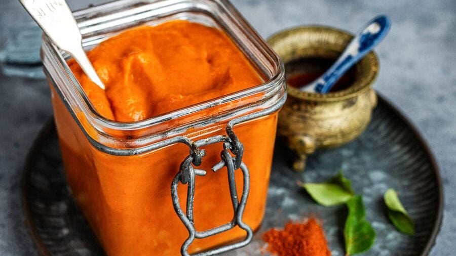 A glass jar filled with bright onion tomato masala sauce sits on a metal tray, with a spoon inside. Nearby are a small brass bowl, a blue spoon, curry leaves, and a pile of red spice powder on a grey surface.