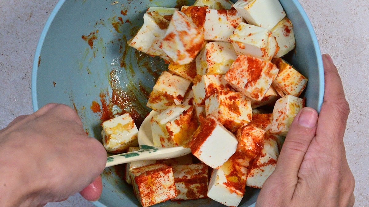 Two hands mix cubed paneer with red and yellow spices in a blue bowl using a spatula.