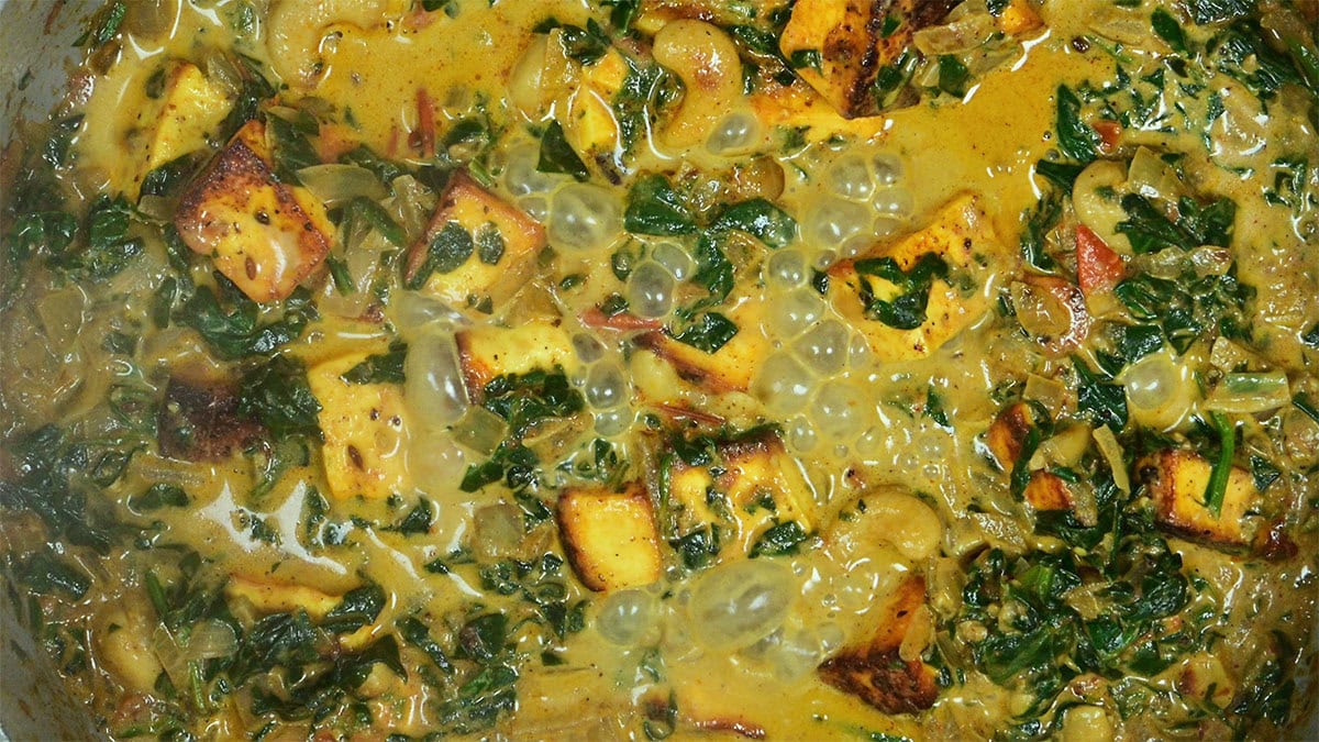 A close-up of a bubbling curry with cubes of paneer, spinach, onions, and cashew nuts in a creamy yellow sauce, showing rich spices and herbs mixed throughout.