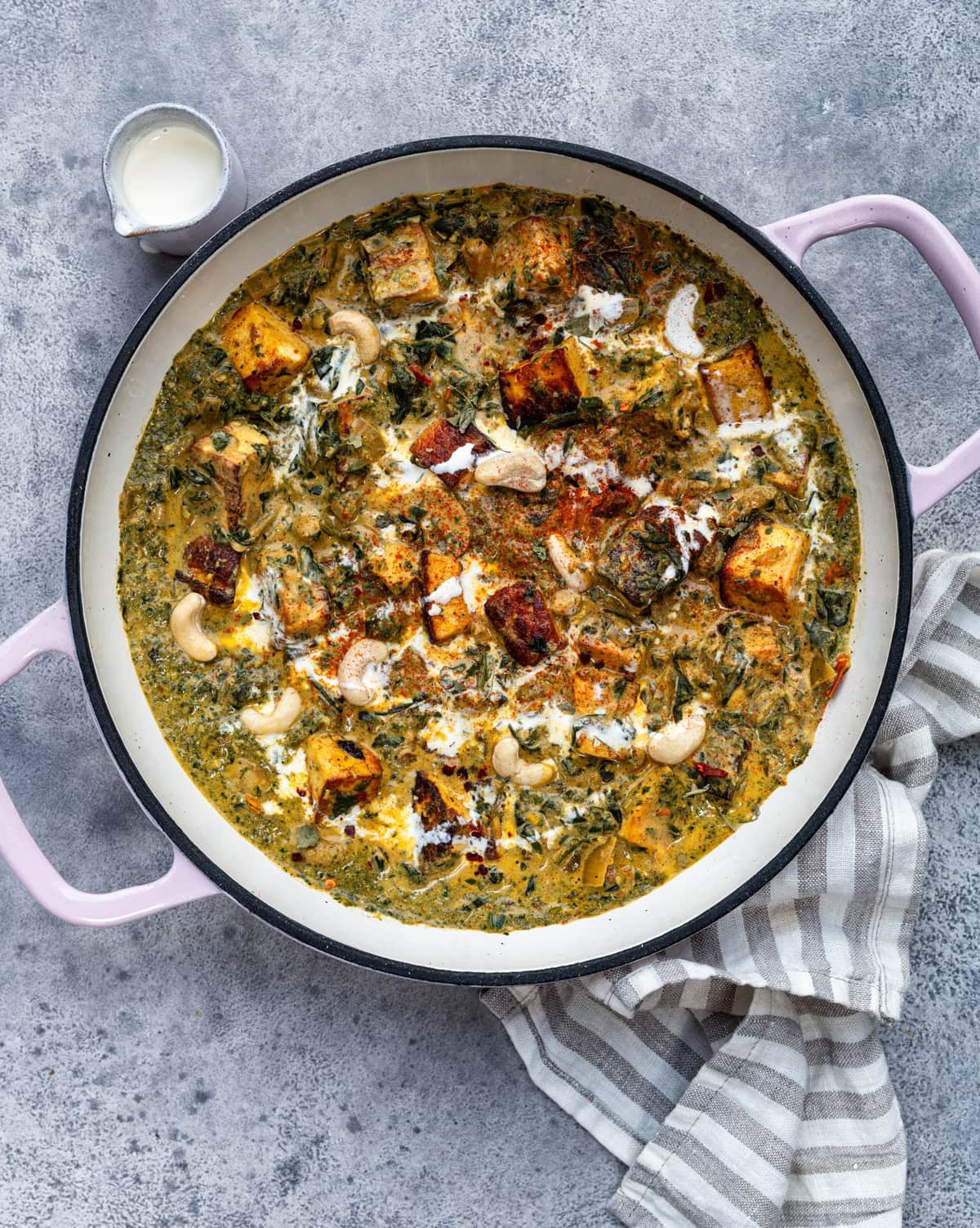 A round pan with handles holds a creamy spinach curry, or saag paneer, topped with paneer cubes, cashews, and a drizzle of cream. A small jug of cream and a striped cloth sit beside the pan on a grey surface.