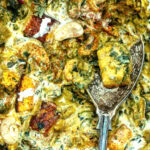 Close-up of a creamy Saag Paneer curry with paneer cubes, cashews, spinach, and spices, with a silver serving spoon resting in the dish. The sauce appears rich and well-seasoned.
