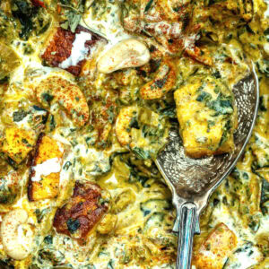 Close-up of a creamy Saag Paneer curry with paneer cubes, cashews, spinach, and spices, with a silver serving spoon resting in the dish. The sauce appears rich and well-seasoned.