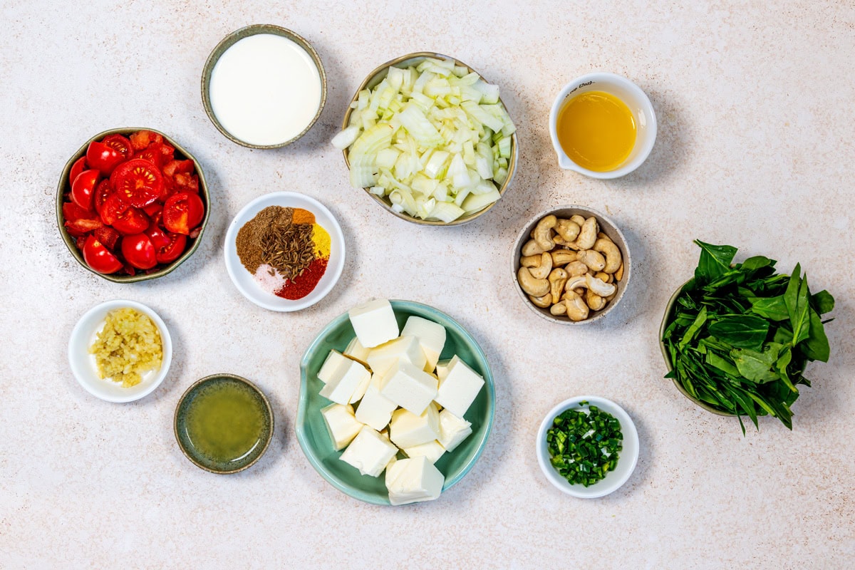 A top view of various ingredients in bowls on a light surface, including chopped onions, cherry tomatoes, cubed paneer, spinach, cashew nuts, chopped green chillies, minced garlic, spices, cream, oil, and lemon juice.