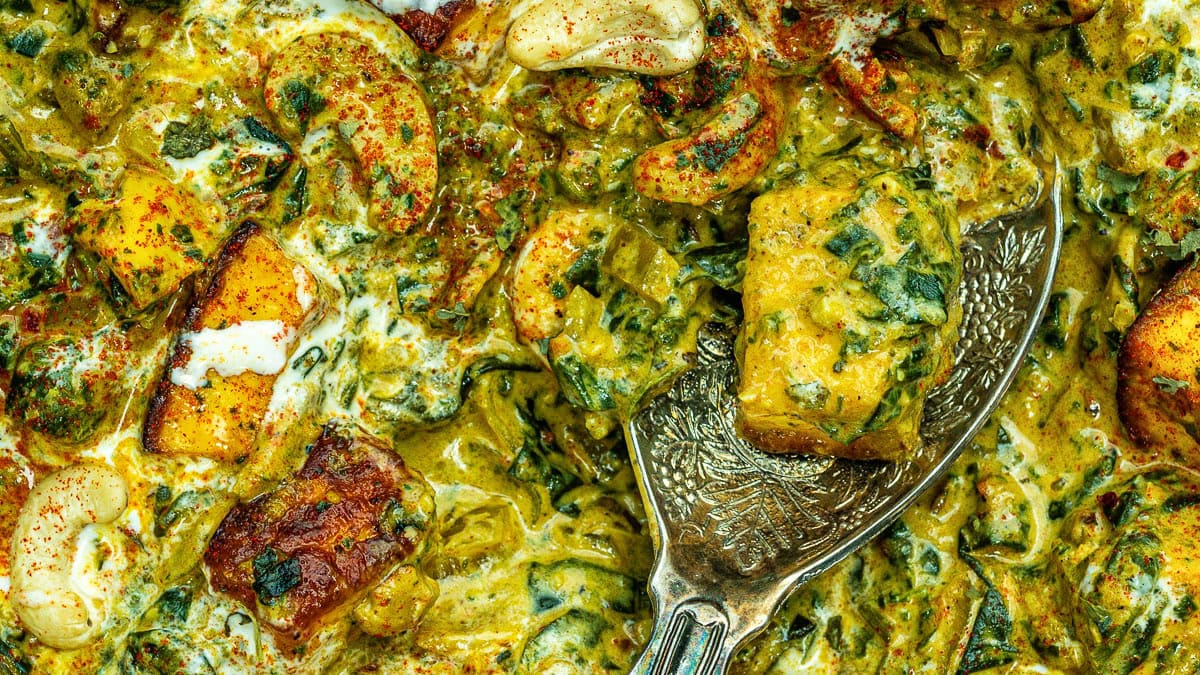 Close-up of a rich, creamy Indian curry with golden paneer cubes, spinach, cashews, and spices, served with a decorative silver spoon. The dish is vibrant with yellow, green, and brown hues.