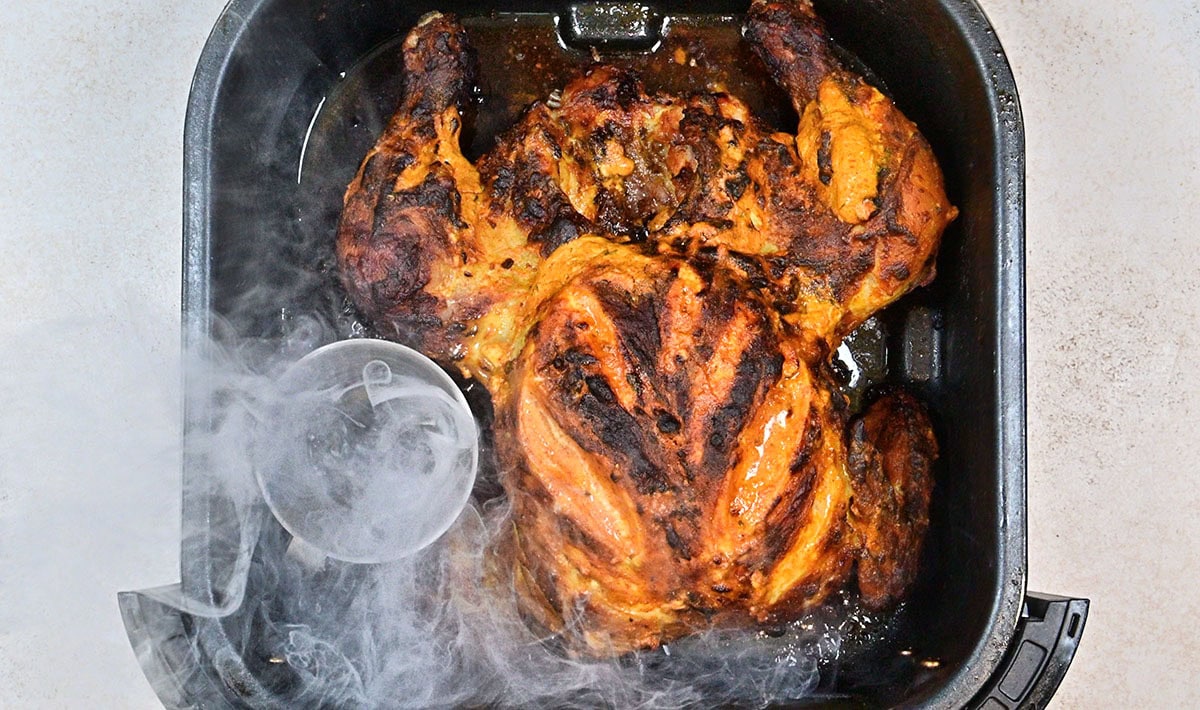 A cooked, tandoori whole chicken with crispy, browned skin sits in an air fryer basket, with steam rising and a small clear container in the corner.
