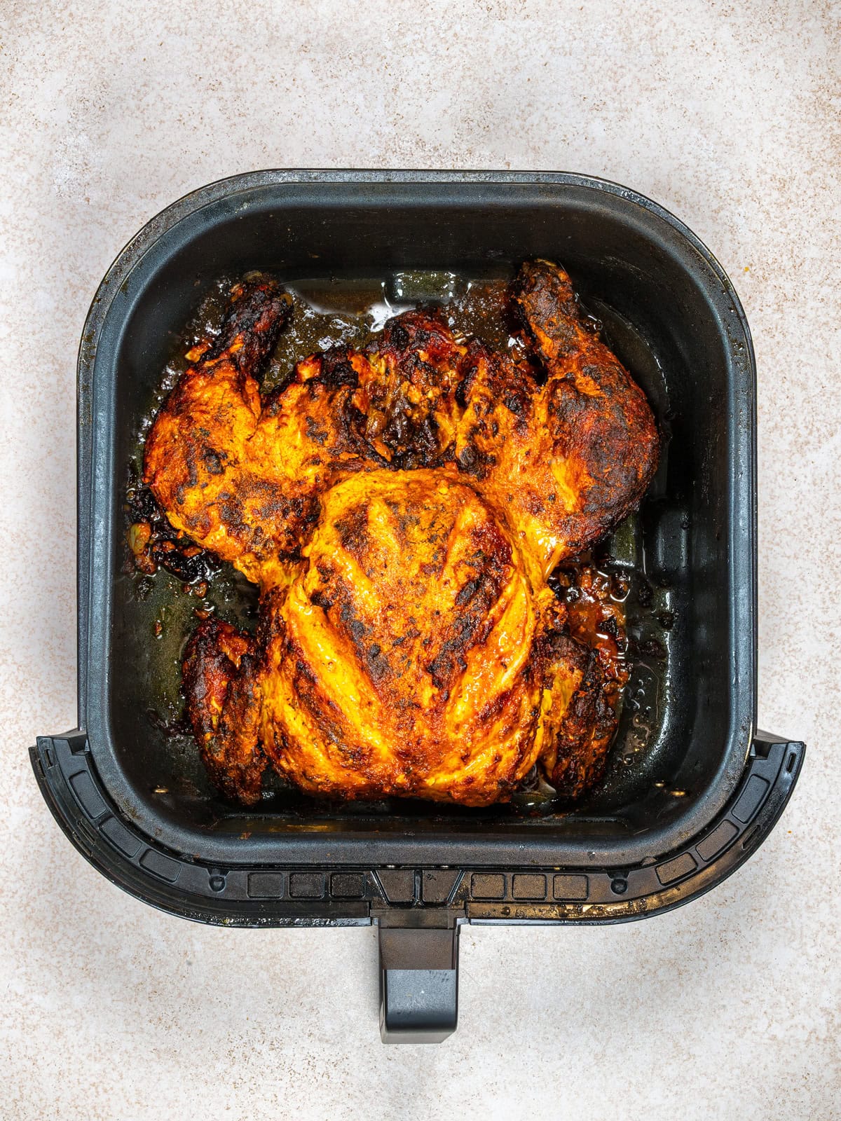 A whole, cooked Tandoori chicken with crispy, seasoned skin sits in the basket of an air fryer, viewed from above on a light-colored surface.