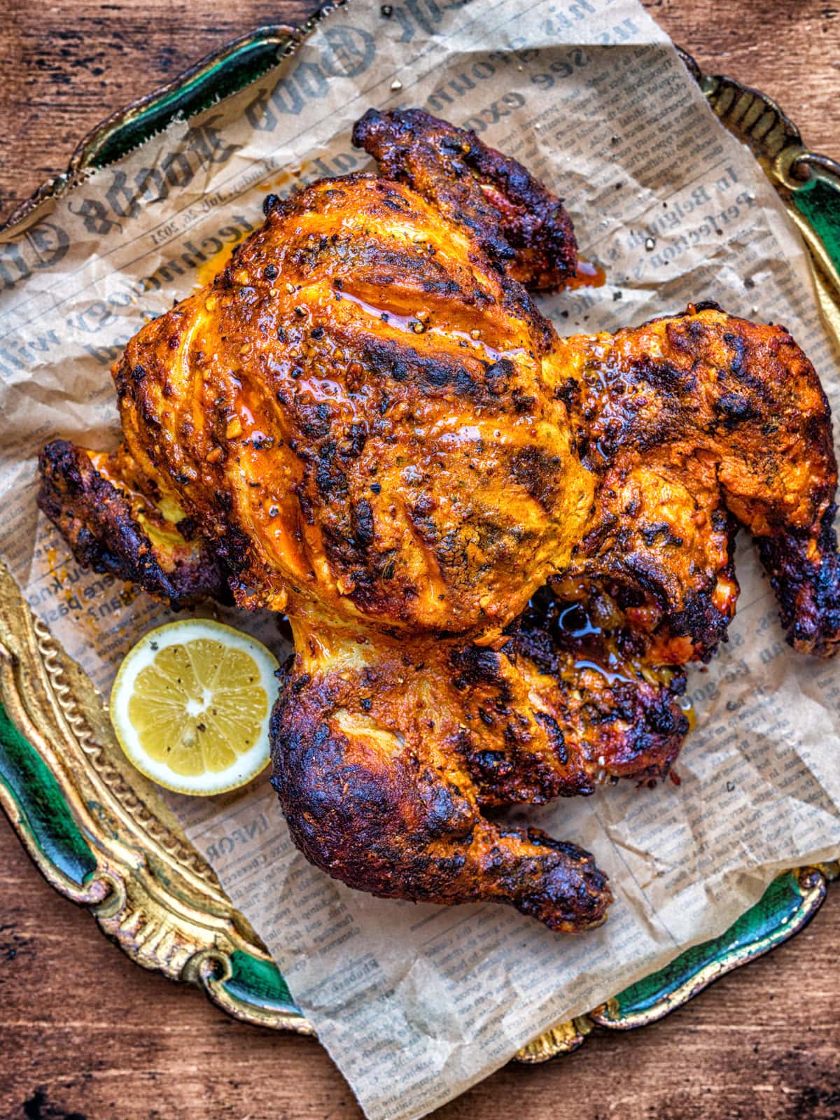 A whole roasted spiced Tandoori chicken with crispy skin sits on newspaper atop a decorative green and gold plate, garnished with a lemon slice on the side.