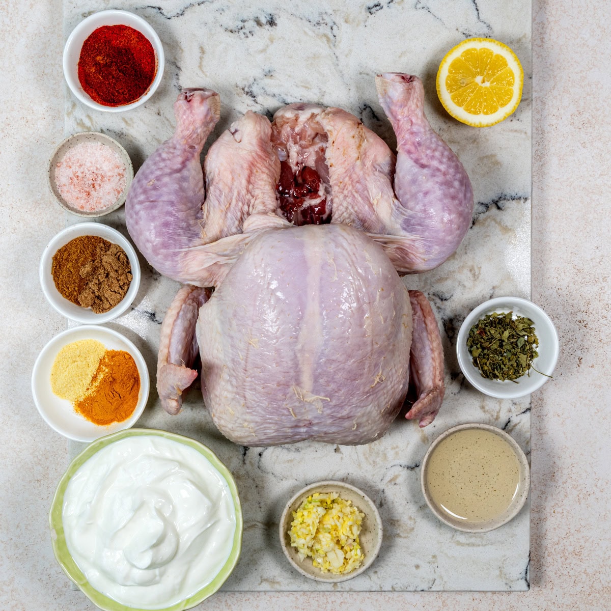 Raw whole chicken on a marble surface, surrounded by bowls of yogurt, spices, ginger-garlic paste, dried herbs, tahini sauce, pink salt, and a half lemon, arranged neatly around the chicken.